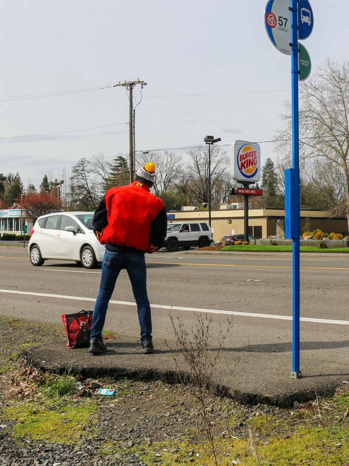 Photo showing a person waiting at a bus stop. The bus stop consists of a broken slab of asphalt perched atop weeds, gravel and trash.