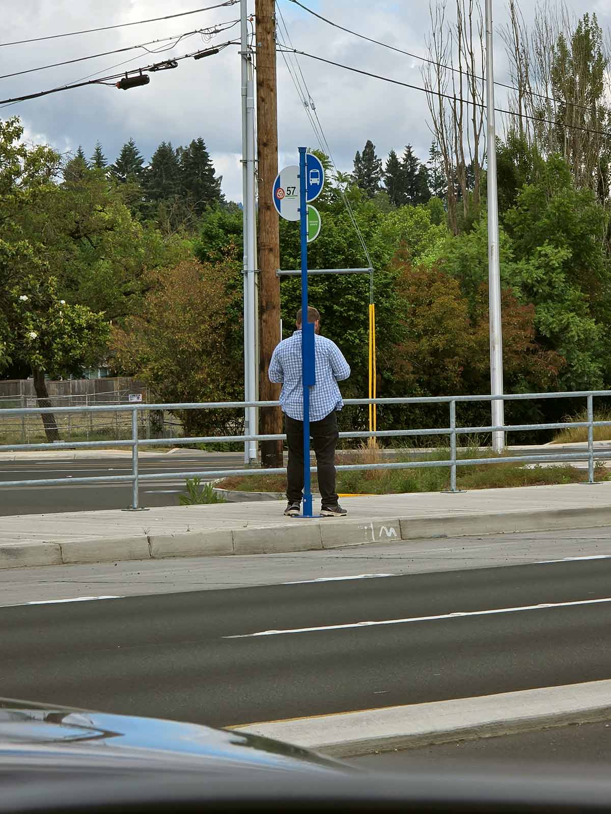 Photo showing a person waiting at a bus stop. The bus stop consists of a broken slab of asphalt perched atop weeds, gravel and trash.